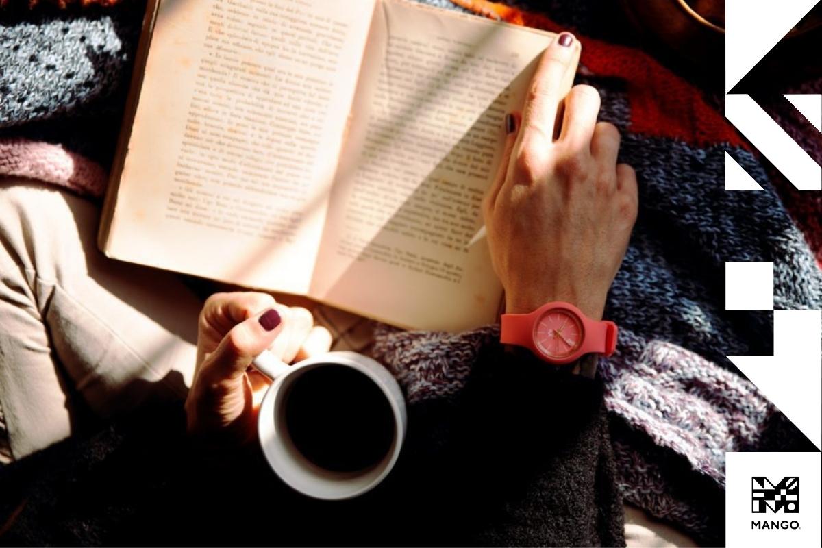 A person sitting down with their legs under a blanket reading a book while drinking coffee