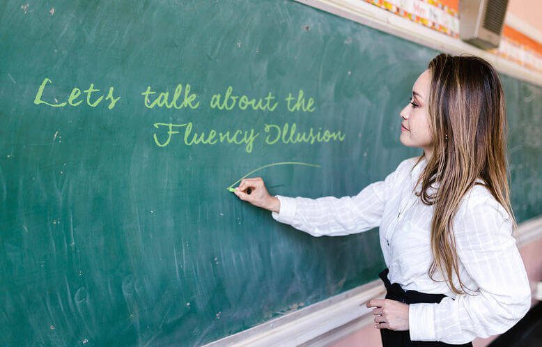 A teacher writing on a chalkboard, she is writing "Let's talk about the fluency illusion"