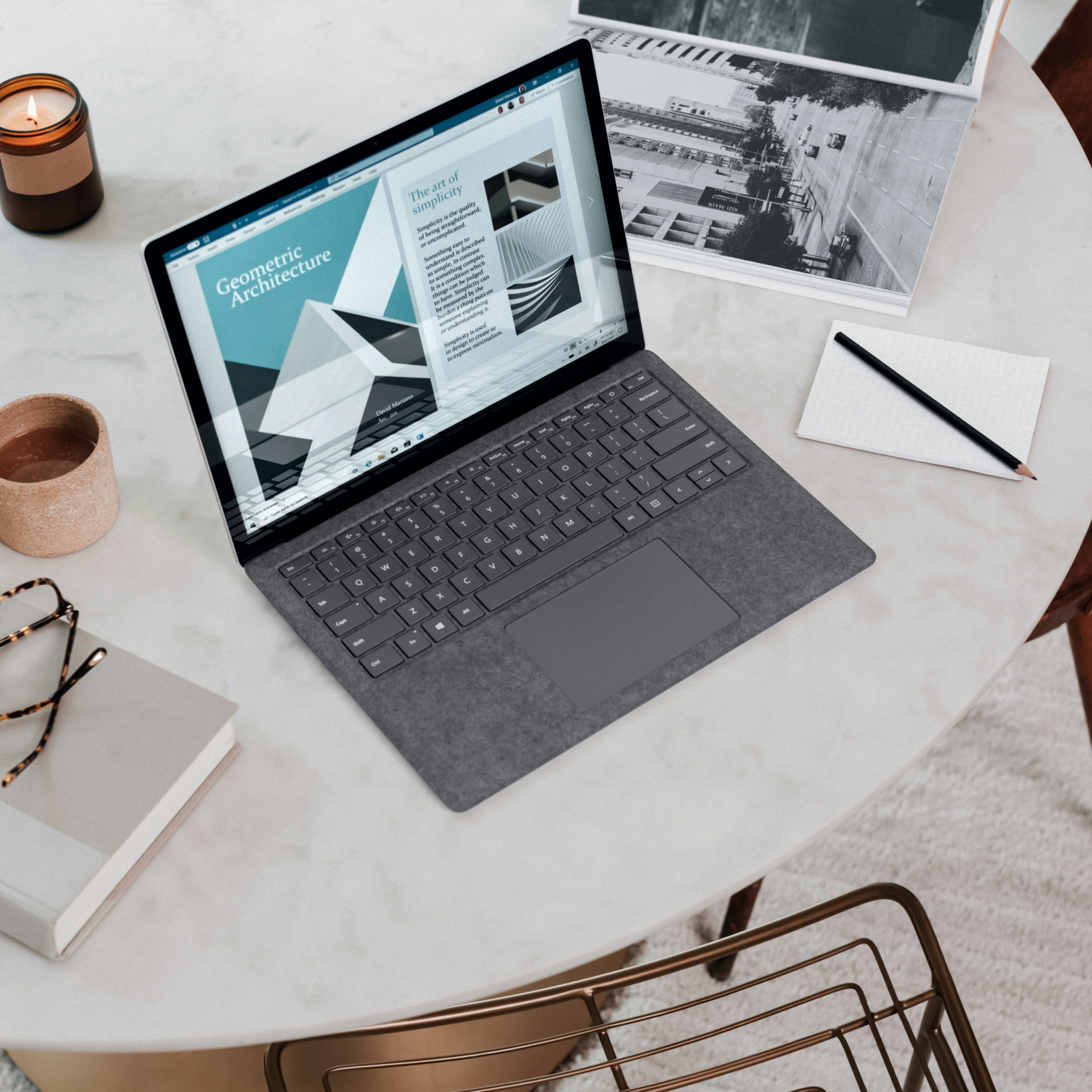 A laptop sits on a white cafe table with some other work materials.